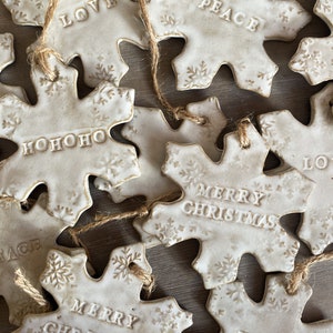 Könnte beinhalten: Eine Sammlung weißer Keramik-Schneeflocken-Ornamente mit verschiedenen Weihnachtsgrüßen, darunter "Love", "Peace", "Merry Christmas", "Hope" und "Ho Ho Ho". Jedes Ornament hat eine Schnur zum Aufhängen.
