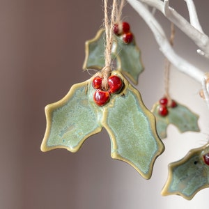 May include: Handmade ceramic holly leaf ornaments with red berry accents, suspended by twine. The leaves are a muted green with a gold trim, ideal for festive decorations. Each ornament is approximately 7.6 cm wide.
