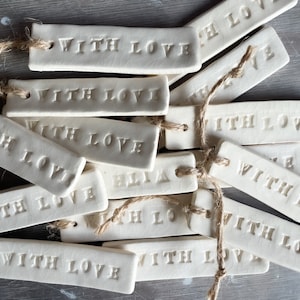 Op de afbeelding: Een verzameling crèmekleurige keramische labels met de tekst "WITH LOVE". Elk rechthoekig label heeft een gaatje en is voorzien van touw. De labels liggen op een grijze ondergrond.