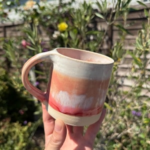 May include: A ceramic mug featuring a gradient glaze that transitions from a creamy off-white at the top to a soft peach, followed by a vibrant pink band near the base. The mug has a sturdy handle and a smooth, unglazed light beige bottom rim.