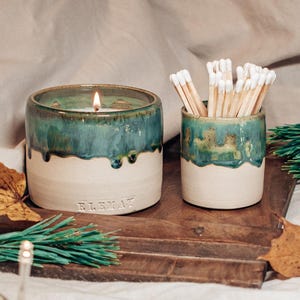 Pode incluir: Um conjunto de velas e porta-fósforos de cerâmica. A vela tem um esmalte verde-azulado sobre uma base creme, com a palavra "ELEMAT" gravada na frente. O porta-fósforos tem um design semelhante e contém fósforos com pontas brancas. O conjunto está em uma bandeja de madeira.