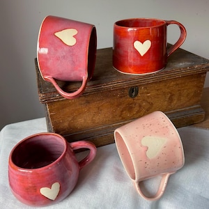 Peut inclure: Quatre mugs en céramique rouges et roses, chacun avec un motif de cœur blanc. Les tasses ont des anses arrondies et sont posées sur un coffre en bois et un tissu blanc. Les tasses varient du rouge foncé au rose clair.