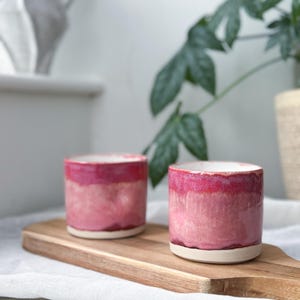May include: Two handmade ceramic cups with a pink ombre glaze. The cups are cylindrical with a white base and a glossy finish. They sit on a wooden cutting board.