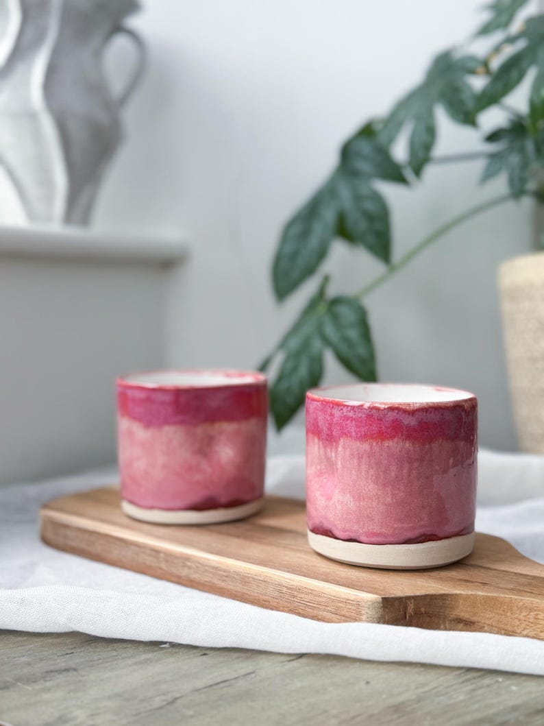 May include: Two handmade ceramic cups with a pink ombre glaze. The cups are cylindrical with a white base and a glossy finish. They sit on a wooden cutting board.