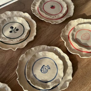 May include: A collection of cream-coloured ceramic plates and smaller bowls with ruffled edges. Each piece features a hand-painted design, including a red tomato, dark blue olives, a pink seashell, and a blue fish, all centred within coloured rings.