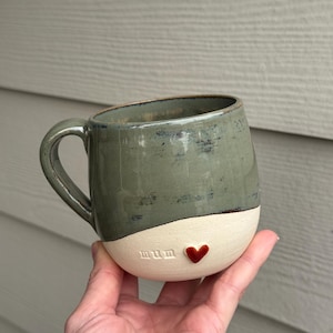 May include: A green ceramic mug with a white bottom and a red heart. The mug says "mum" on the bottom.