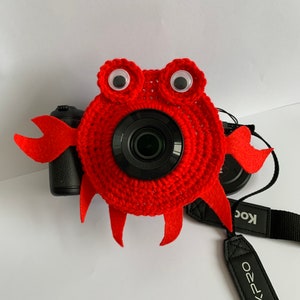 May include: A red crocheted camera cover in the shape of a crab, with googly eyes and felt claws. The cover is on a black camera with a lens. A black camera strap with the text "PIXPRO" is visible.