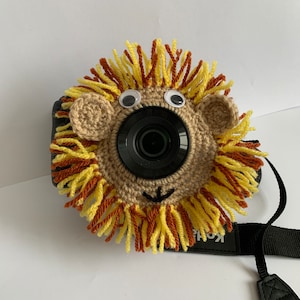 May include: A crocheted camera cover in the shape of a lion's face. The cover is made with tan yarn and has a yellow and brown yarn mane, googly eyes, and small tan ears. The camera strap is black.