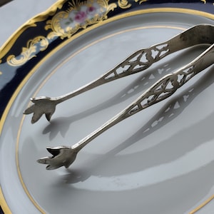 May include: Silver sugar tongs with an ornate, openwork design. The tongs are resting on a white plate with a blue and gold floral pattern.