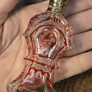 May include: A clear glass perfume bottle with red decorative accents and a gold-colored cap. The bottle features an ornate design with red etched patterns and a detailed stopper. The bottle is held in a hand.