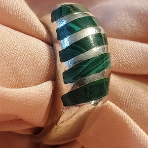 May include: A silver ring with alternating bands of polished silver and vibrant green malachite. The ring's design features a series of horizontal stripes, creating a bold and eye-catching pattern. The ring is displayed on a soft, peach-colored fabric.