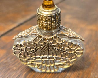 May include: A small, clear glass perfume bottle with an ornate gold-colored filigree design. The bottle has a rounded shape and a decorative gold-colored stopper with a brown gem on top. The bottle is likely vintage or antique.