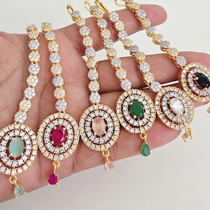 Może przedstawiać: Sześć złotych ozdób do włosów tikka z kamieniami centralnymi w różnych kolorach, w tym zielonym, różowym, czerwonym i niebieskim. Każdy element ma okrągły kształt z wiszącym kamieniem w kształcie łzy. Ozdoby są ozdobione małymi, błyszczącymi kryształami.