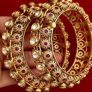 May include: Gold-toned bangles with intricate floral and geometric designs. The bangles feature red and clear gemstones, with green accents. The jewelry is displayed against a red background, showcasing the ornate details.