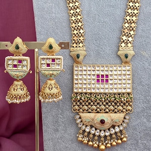 May include: Gold-toned jewelry set featuring a long necklace and matching earrings. The necklace has a rectangular pendant with a grid pattern and red accents. The earrings are jhumka style with green and red details. The set is displayed on a gray stand.