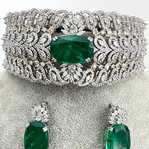 Op de afbeelding: Een zilveren choker ketting met een grote smaragdgroene steen omringd door kleinere heldere stenen. Passende oorbellen met smaragdgroene stenen en heldere stenen.