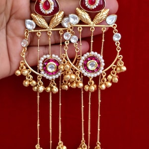 Puede incluir: Pendientes colgantes dorados con un diseño rojo y blanco. Los pendientes presentan una piedra roja triangular en la parte superior, con una piedra blanca central y un borde perlado. El diseño incluye cadenas colgantes con pequeñas cuentas doradas.