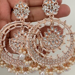 May include: A pair of large, round, gold-tone earrings with white pearl accents. The earrings feature intricate designs with multiple layers of sparkling rhinestones.