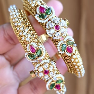 May include: Two gold bangles with detailed patterns. Each bangle has a floral design with red, green, and clear gemstones, framed by small white beads. The bangles have a textured surface and a clasp closure.