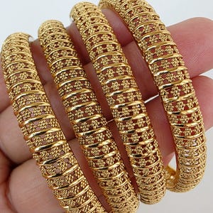 Op de afbeelding: Vier goudkleurige armbanden met ingewikkelde bloemen- en stippenpatronen. De armbanden worden in een hand gehouden, waardoor hun gedetailleerde ontwerp en gepolijste afwerking worden benadrukt. De sieraden hebben een heldere, warme goudkleur.