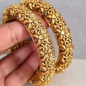 May include: Two gold bangles with detailed floral patterns. The bangles are thick and feature an embossed design of flowers and leaves. The jewellery is displayed against a neutral background.