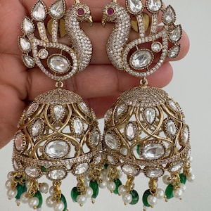 May include: A pair of gold-coloured peacock earrings. The earrings feature a peacock design with clear, oval-shaped stones and small red stones. The earrings have a bell-shaped bottom with clear stones and green and white bead accents.
