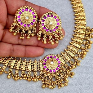 Könnte beinhalten: Ein goldfarbenes Schmuckset mit Halskette und passenden Ohrringen. Die Ohrringe sind rund mit einem zentralen Design und einem lila Rand. Die Halskette hat ein ähnliches Design mit einem zentralen Anhänger und hängenden goldenen Perlen.