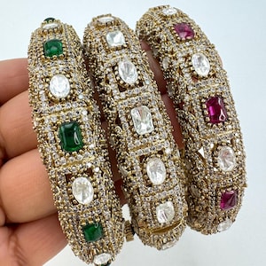 Peut inclure: Trois bracelets en or avec des motifs complexes. Les bracelets sont ornés de pierres claires et colorées. Les pierres sont disposées en forme de carré. Les bracelets sont empilés les uns sur les autres.
