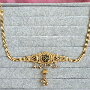 May include: A gold waist chain with detailed patterns. The central section has a dark green and red design, with small red and green gems. Dangling bells and beads provide movement to the piece.