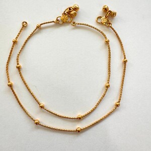 K&ouml;nnte beinhalten: Ein Paar goldene Fu&szlig;kettchen mit kleinen runden Perlen und dekorativen Anh&auml;ngern. Die Fu&szlig;kettchen bestehen aus einer zarten Kette.