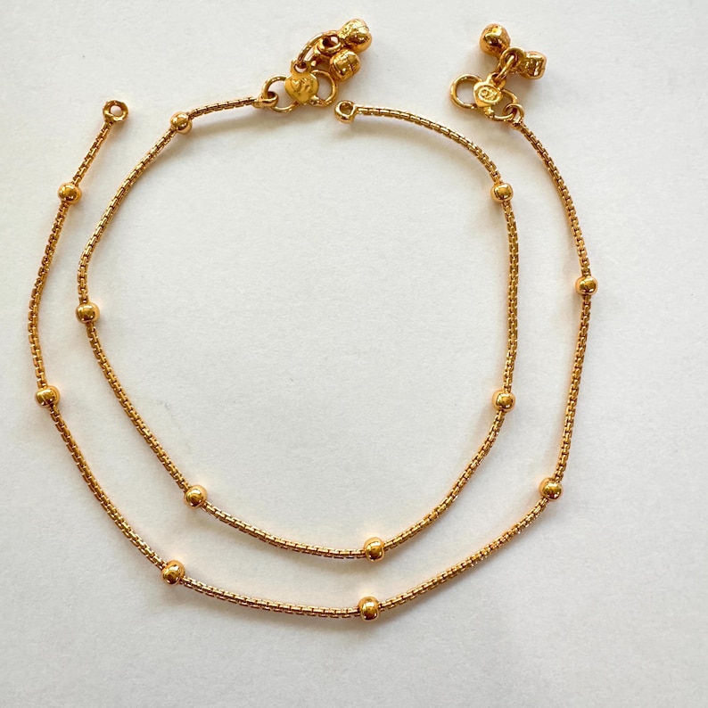 K&ouml;nnte beinhalten: Ein Paar goldene Fu&szlig;kettchen mit kleinen runden Perlen und dekorativen Anh&auml;ngern. Die Fu&szlig;kettchen bestehen aus einer zarten Kette.