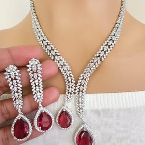 Könnte beinhalten: Ein schillerndes Schmuckset mit Halskette und passenden Ohrringen. Die Halskette und die Ohrringe sind mit klaren, funkelnden Steinen und großen, tropfenförmigen, rubinroten Edelsteinen verziert. Die Halskette hat ein Y-förmiges Design, und die Ohrringe sind lang.