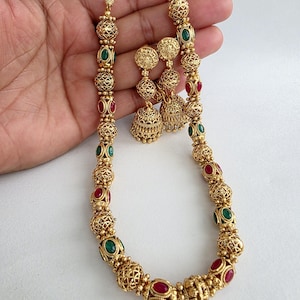 May include: A gold-toned necklace and matching earrings set. The necklace features alternating red and green gemstone accents within ornate, filigree-style beads. The earrings have a similar design, with dangling bell-shaped pendants. The jewelry is displayed against a white background.
