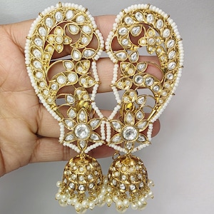 Op de afbeelding: Gouden oorbellen met een ingewikkeld filigraanontwerp en heldere edelsteenaccenten. De oorbellen hebben een hartvormig ontwerp met een rand van kleine witte kralen. Het onderste deel heeft een belvorm met hangende witte parels.