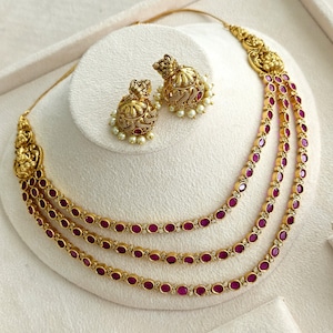 May include: A gold-toned jewelry set consisting of a three-strand necklace and matching jhumka earrings. The necklace features oval-shaped magenta stones set in gold frames, accented with ornate gold motifs. The earrings have a bell-shaped design.