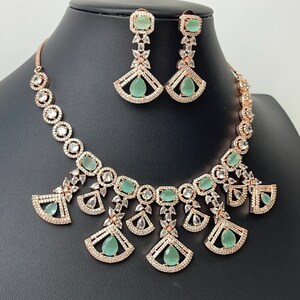 Pode incluir: Um conjunto de colar e brincos de ouro rosa com pedras transparentes e verde claro. O colar apresenta uma corrente delicada com um pingente de v&aacute;rios desenhos em forma de leque com pedras transparentes e pedras verde claro. Os brincos s&atilde;o brincos pendentes com uma pedra verde claro e pedras transparentes.