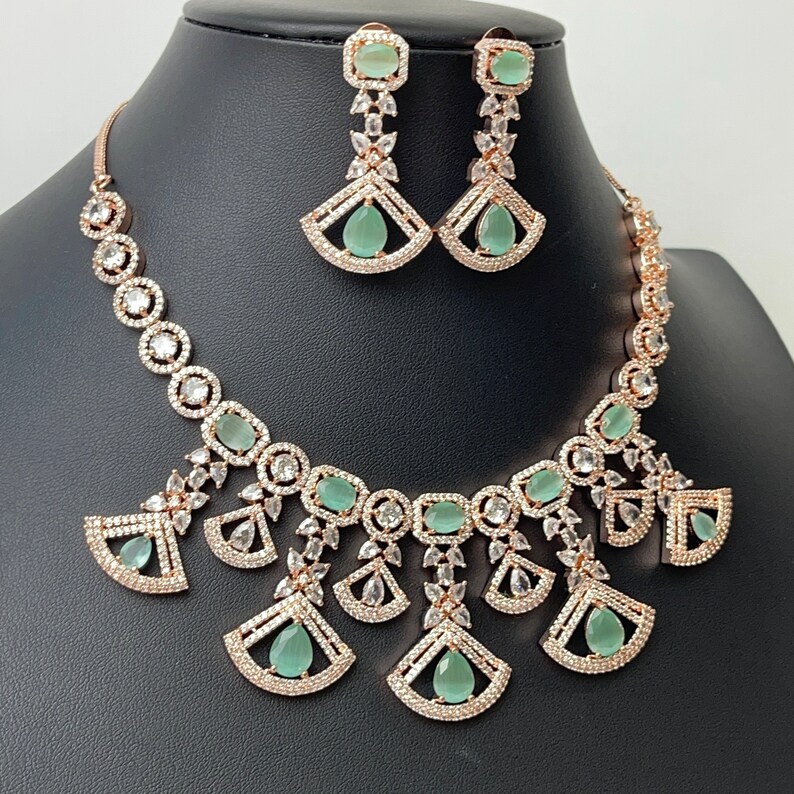 Pode incluir: Um conjunto de colar e brincos de ouro rosa com pedras transparentes e verde claro. O colar apresenta uma corrente delicada com um pingente de v&aacute;rios desenhos em forma de leque com pedras transparentes e pedras verde claro. Os brincos s&atilde;o brincos pendentes com uma pedra verde claro e pedras transparentes.