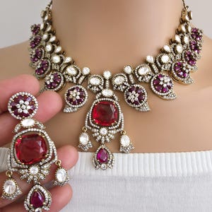 May include: A vintage-style necklace and earring set. The jewelry features large, square, and teardrop-shaped red gemstones, surrounded by smaller clear and purple stones. The metal has a gold tone.