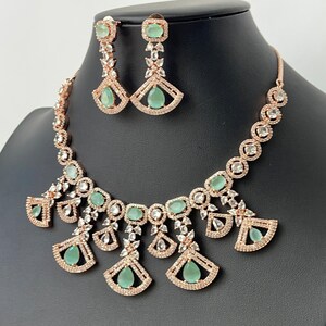 Pode incluir: Um conjunto de colar e brincos de ouro rosa com pedras transparentes e verde claro. O colar apresenta um design de pingente com v&aacute;rias pedras em forma de l&aacute;grima e um design em forma de leque.