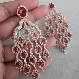 Könnte beinhalten: Ein Paar roségoldene Chandelier-Ohrringe. Jeder Ohrring hat einen großen, tropfenförmigen, roten Edelstein an der Basis, mit kleineren roten Tropfen-Edelsteinen, die nach oben kaskadieren. Die Ohrringe sind mit klaren, funkelnden Akzenten verziert.