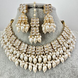 May include: A gold and white bridal necklace set with pearl accents. The necklace features a layered design with multiple rows of pearls and a central section of sparkling stones. The earrings are similarly designed with a smaller version of the necklace's central section.