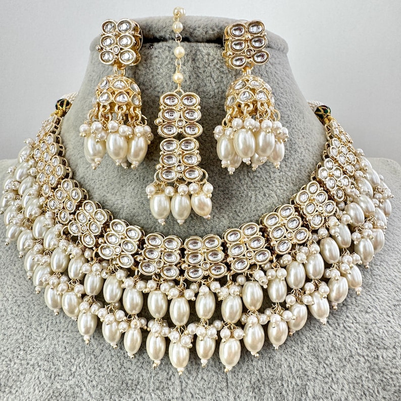 May include: A gold and white bridal necklace set with pearl accents. The necklace features a layered design with multiple rows of pearls and a central section of sparkling stones. The earrings are similarly designed with a smaller version of the necklace's central section.