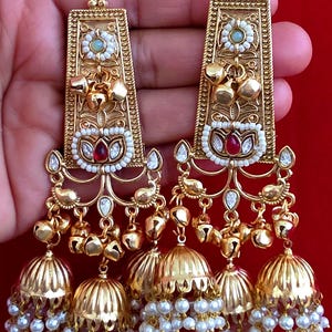 May include: A pair of gold-toned dangle earrings featuring an elongated rectangular top section with a floral motif, a central red stone surrounded by white beads, and three bell-shaped jhumka drops at the base adorned with dangling white pearls.