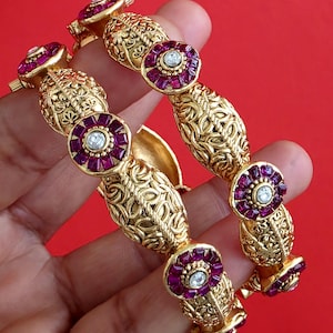 May include: Two gold-tone metal bangles featuring intricate filigree patterns and alternating circular motifs. Each circular element is set with deep red square-cut stones surrounding a single clear central stone, creating a repeating floral design.
