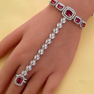 May include: A silver hand chain bracelet with a ring, featuring square red gemstones and small clear stones. The bracelet connects to the ring with a delicate chain of clear stones. The bracelet is on a light yellow background.