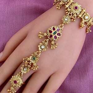 May include: A gold-toned hand harness featuring a floral-linked chain connecting a wrist bracelet to a finger ring. The design incorporates intricate metalwork, small dangling beads, and inlaid red and green stones arranged in circular patterns.