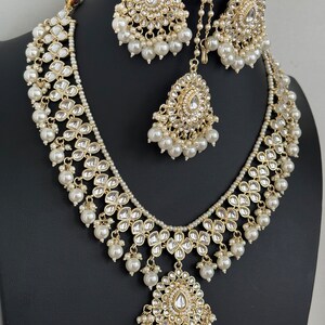 May include: A gold necklace set with a large pendant, featuring intricate floral designs and white pearls. The set includes matching earrings and a maang tikka.