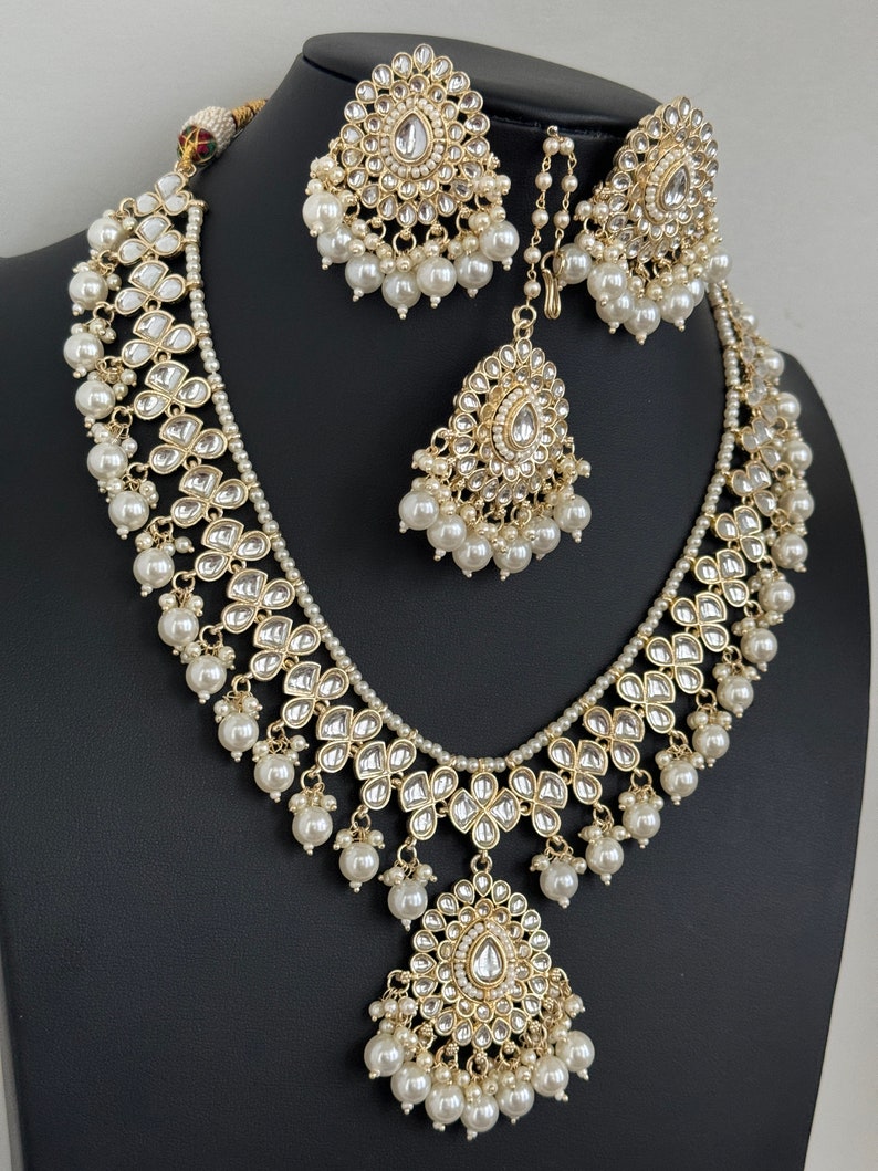 May include: A gold necklace set with a large pendant, featuring intricate floral designs and white pearls. The set includes matching earrings and a maang tikka.