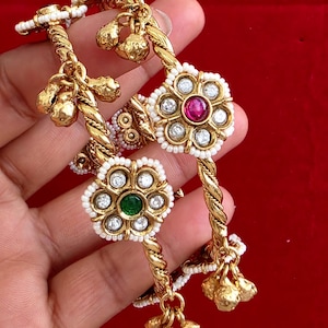 May include: Two gold-toned metal bangles featuring a twisted rope design. Each bangle is adorned with a central flower-shaped motif set with white stones and a single red or green stone, surrounded by small white beads and dangling gold bells.