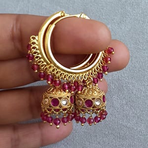 May include: Gold-toned hoop earrings with dangling ruby-colored beads and bell-shaped dangles. The earrings have intricate detailing and are held up by a person's fingers against a gray background. The earrings are a type of jewelry.